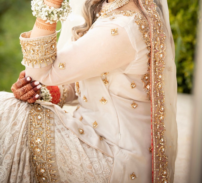 May include: A white bridal outfit with intricate gold embroidery and a matching veil. The outfit features a long-sleeved top and a flowing skirt.