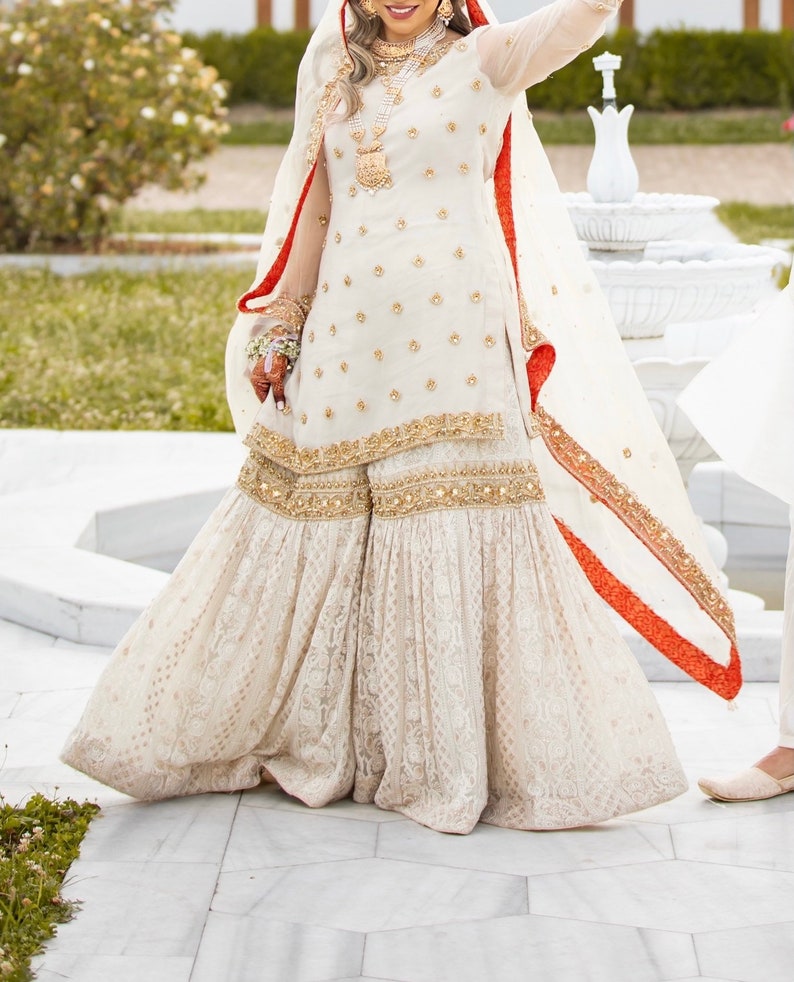 May include: A white bridal outfit with gold embroidery and a red trim. The outfit includes a long-sleeved top and a flared skirt.