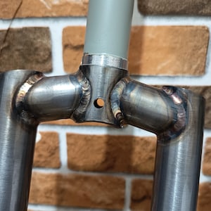 Pode incluir: Uma coroa de garfo de bicicleta em aço bruto sem pintura, com soldaduras visíveis e tons de calor que ligam as duas pernas do garfo ao tubo de direção central. O tubo de direção é pintado de cinzento claro e tem um pequeno orifício circular.