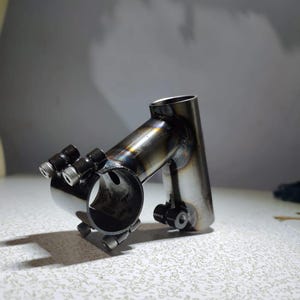 May include: A black metal bicycle stem with a silver clamp. The stem is designed to attach a handlebar to a bicycle frame.