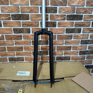 Puede incluir: Horquilla de bicicleta con un vástago plateado y patas negras. La horquilla está diseñada para una bicicleta y está hecha de metal. El fondo es una pared de ladrillos.