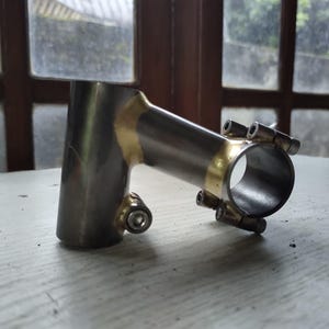 May include: A silver-colored bicycle stem with gold-colored accents. The stem has a cylindrical shape with multiple bolt holes for attachment. The product is designed to connect the handlebars to the fork of a bicycle.