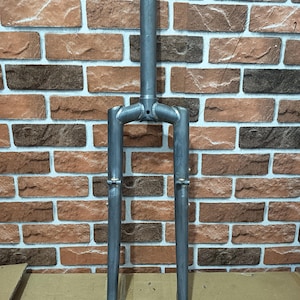 Puede incluir: Horquilla de bicicleta plateada con dos patas paralelas y un vástago central. La horquilla tiene una construcción soldada y está diseñada para fijarse a un cuadro de bicicleta.