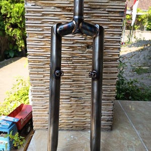 May include: A raw steel bicycle fork featuring a cylindrical steerer tube, two straight fork blades, and visible heat-treated weld marks at the crown. The fork includes two mounting bosses on the inner side of each blade for accessory attachment.