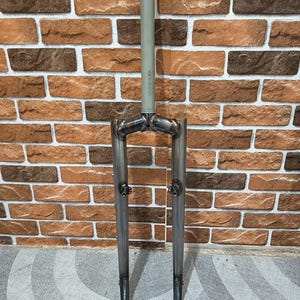 May include: A silver bicycle fork with a long, vertical tube and two parallel, curved legs. The fork is made of metal and has a welded construction. The background is a brick wall.