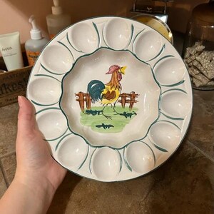 May include: A round white ceramic egg serving platter featuring a scalloped edge and twelve oval indentations. The center of the platter displays a hand-painted illustration of a rooster standing in front of a brown fence on green grass.