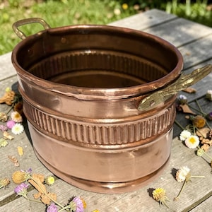 May include: A copper-colored metal bucket with two brass handles. The bucket has a decorative band of vertical lines around the middle. The bucket is sitting on a wooden surface with dried flowers.