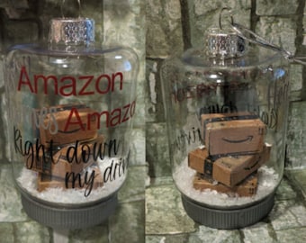 May include: A clear glass Christmas ornament with a silver top and a white base. Inside the ornament are several miniature cardboard boxes with the Amazon logo. The ornament has the text "Amazon loves Amazon right down to my drive" written on it.