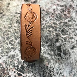 May include: A tan leather bracelet with a floral design. The design features roses and leaves.