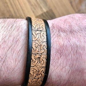 Könnte beinhalten: Ein schwarzes Lederarmband mit einer braunen Ledereinlage, die ein keltisches Knotendesign zeigt.