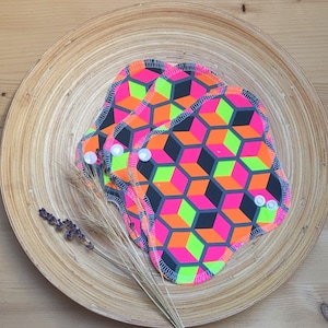 Könnte beinhalten: Drei wiederverwendbare Stoff-Menstruationseinlagen mit einem geometrischen Muster aus rosa, orange, grün und schwarzen Quadraten. Die Einlagen sind gefaltet und übereinander gestapelt.