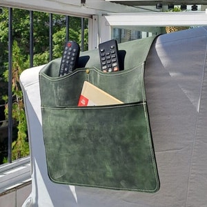 Könnte beinhalten: Ein grüner Leder-Fernbedienungshalter mit zwei Taschen und weißen Nähten. Der Halter ist an einem weißen Stuhl befestigt und enthält zwei Fernbedienungen und ein Buch.