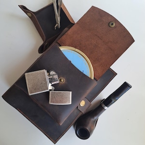 May include: A brown leather tobacco pouch with a silver lighter, a pipe, and a tin of tobacco. The pouch is open, revealing the contents. A small pair of silver tools is also visible.