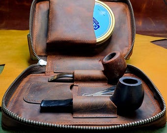 May include: A brown leather pipe case with a zipper closure. The case is open and contains two pipes, a small tin, and a pipe tool. The case is made of distressed leather and has a vintage look.