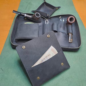 Könnte beinhalten: Eine schwarze Ledertasche mit Reißverschluss. Die Tasche hat mehrere Fächer und Taschen zum Verstauen verschiedener Gegenstände. Die Tasche ist geöffnet und zeigt eine Pfeife, eine kleine Tasche und ein kleines Stück Papier mit dem Text "Karelias and Sons".