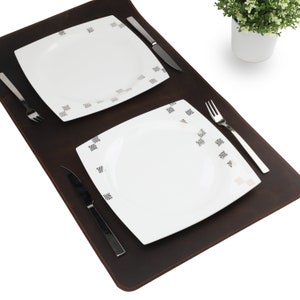 May include: Two white ceramic dinner plates with a black and white geometric pattern on a brown leather placemat. Two silver forks and two silver knives are placed on the placemat.