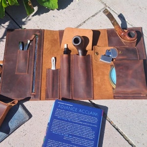 May include: A brown leather roll-up case with multiple pockets and compartments for storing smoking pipes, tools, and accessories. The case is open and laid flat on a concrete surface. A book titled "Advance Acclaim" is visible in the foreground.
