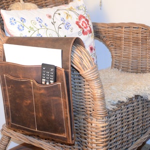 May include: A brown leather chair arm organizer with two pockets. The organizer has a remote control and a piece of paper in the pockets. The organizer is attached to a wicker chair.