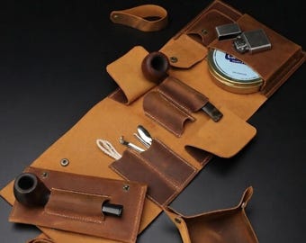 May include: A tan leather tobacco pipe roll-up organizer featuring multiple pockets, a dark brown wooden pipe, a metal lighter, a round tin, and metal pipe tools. The set includes a matching leather tray and a leather strap for secure closure.