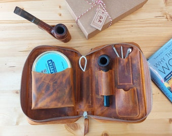 Puede incluir: Una bolsa de tabaco de cuero marrón con cierre de cremallera. La bolsa tiene varios bolsillos y compartimentos para guardar tabaco, una pipa y otros accesorios. La bolsa está abierta y el interior es visible, revelando una pipa, una lata de tabaco y un tampón para pipa.