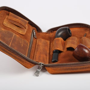 Può includere: Un astuccio per pipe in pelle marrone con chiusura a zip. L'astuccio è aperto e contiene due pipe, un tampone e uno strumento di pulizia.