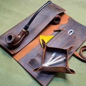 Pode incluir: Um estojo de couro marrom para cachimbo de tabaco com um cachimbo, uma pequena bandeja com ferramentas e uma bolsa. O cachimbo é feito de madeira e tem um bocal preto. O estojo está aberto, revelando um interior marrom.