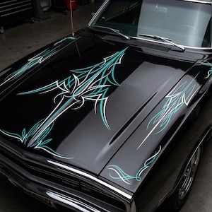 May include: Close-up of a black car hood with custom pinstriping. The design features teal and white lines and shapes, creating a dynamic and artistic pattern. The hood is glossy, reflecting the surrounding environment.