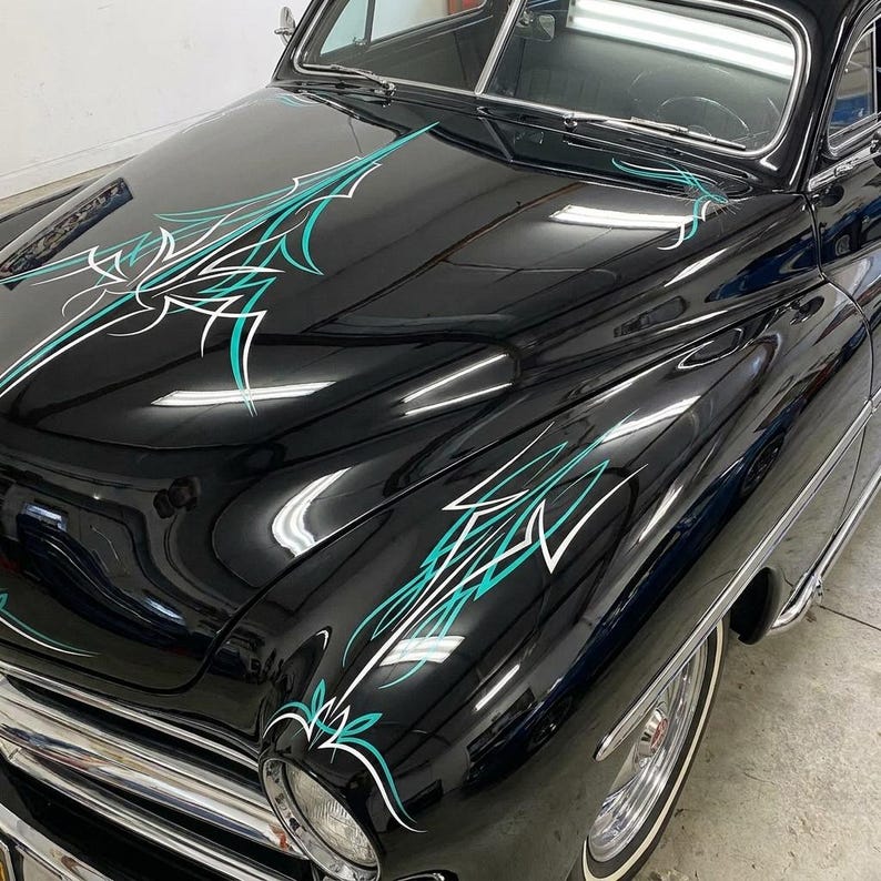 May include: A classic black car with custom teal and white pinstriping on the hood and fenders. The car has chrome accents, a white wall tire, and a vintage design. The paint is glossy, reflecting the surrounding environment.