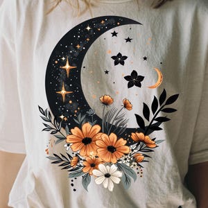 May include: Cream-colored t-shirt featuring a celestial design. A black crescent moon is adorned with stars and a constellation. Orange and white flowers and foliage are arranged below the moon, creating a whimsical, nature-inspired graphic.