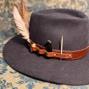May include: A dark grey fedora hat with a brown leather band and decorative feathers. The hat features a small black stone and a wooden toothpick. The hat is a stylish accessory.