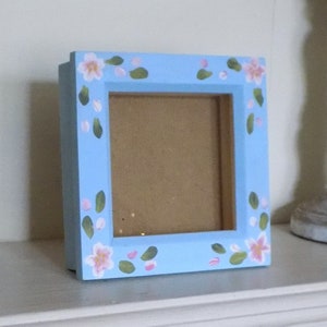 May include: A square wooden picture frame painted in a light blue colour. The frame features a hand-painted floral pattern consisting of small pink blossoms and green leaves distributed evenly around the border of the frame's front face.