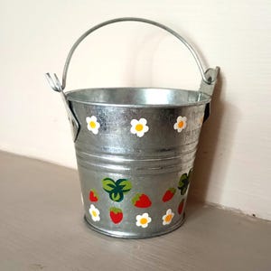 May include: A small galvanised metal bucket with a silver finish and a swinging handle. The exterior is decorated with a repeating pattern of small white flowers with yellow centres, red strawberries, and green leaves painted around the body.