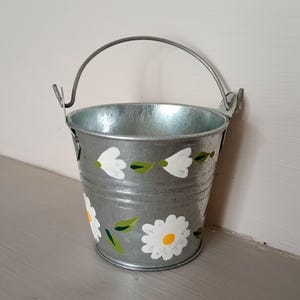 May include: A small, silver-coloured metal bucket featuring a hand-painted floral design. The exterior is decorated with white daisy-like flowers with yellow centres and green leaves arranged in a horizontal pattern around the cylindrical body.