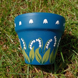 May include: A hand-painted, blue ceramic flower pot. The pot is decorated with white lily-of-the-valley flowers and green leaves. The pot is sitting on a bed of green grass.