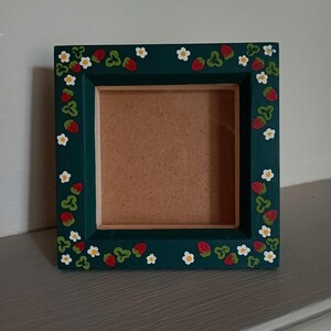 May include: A square picture frame with a dark green painted border featuring a hand-painted pattern of small red strawberries, green leaves, and white flowers with yellow centres. The centre of the frame is a plain, light brown square backing.