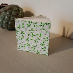 May include: A miniature open book with a white cover featuring a green leaf design. The book sits on a light surface, with a green decorative item in the background. The pages are visible, indicating a handmade journal or notebook.