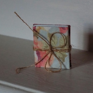 May include: A small, fabric-covered book with a floral pattern in shades of pink, green, and blue. The book is tied with twine and has a rustic, handmade aesthetic.