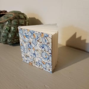 May include: A small, open notebook featuring a floral design of blue flowers on a white backdrop. The pages are white, and the notebook is standing upright. The notebook is on a light grey surface.