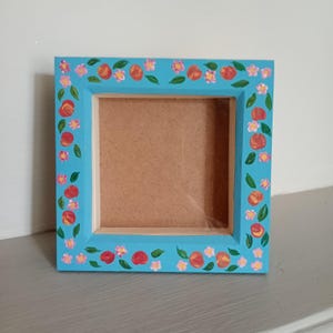 May include: A square wooden picture frame painted in a bright turquoise blue colour. The frame features a hand-painted pattern of small orange peaches, pink flowers, and green leaves distributed evenly around the entire border of the frame.