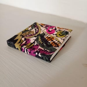 May include: A small, square notebook with a colourful paisley pattern in pink, green, gold, and orange on a cream background. The edges are black. The notebook is resting on a light grey surface.