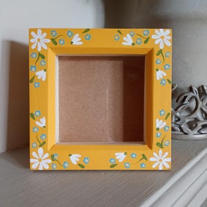 May include: Square yellow picture frame with white daisy and blue forget-me-not floral design. The frame is painted with a bright yellow colour and has a natural wood backing. The frame is approximately 15 cm square.