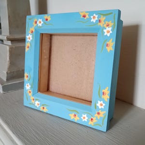 May include: A square, light blue picture frame decorated with hand-painted yellow and white daffodil flowers. The frame is made of wood and has a natural wood backing. The frame is displayed on a grey surface.