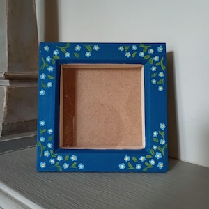May include: Square blue picture frame with a floral design. The frame is painted a deep blue and decorated with small blue flowers and green leaves. The inside of the frame is a light brown colour.