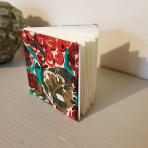 May include: A small notebook with a floral fabric cover. The cover displays a pattern of red, teal, and brown flowers. The pages are white and fanned out, showing the notebook's thickness. The notebook is on a light grey surface.