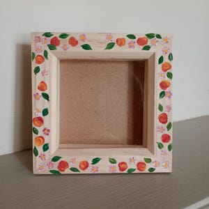 May include: A square wooden picture frame with a light natural finish. The frame border is hand-painted with a repeating pattern of orange peaches, small purple flowers, and green leaves arranged around the entire perimeter of the frame.