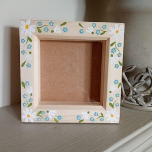 May include: Square wooden picture frame with hand-painted floral decoration. The frame is made of light-coloured wood, adorned with white daisies and small blue flowers. The flowers are arranged around the edges.