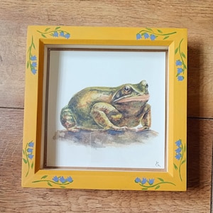 May include: A framed watercolour painting of a frog. The frog is depicted in shades of green, brown, and tan, resting on a rock. The frame is yellow with blue floral accents. The artwork is a square shape.