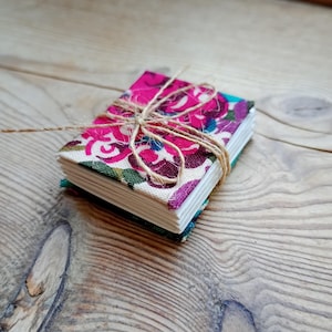 May include: A miniature notebook with a floral pattern, bound with twine. The cover displays a bright pink and purple floral design on a cream backdrop. The notebook rests on a teal and green patterned fabric.
