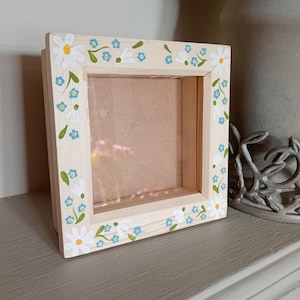 May include: A square wooden picture frame with a natural finish, adorned with hand-painted white daisies and blue forget-me-nots. The frame features a clear front and a light brown backing, ideal for displaying photos or artwork.