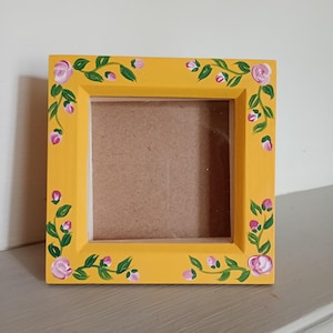 May include: A square picture frame painted in a bright yellow colour. The frame features hand-painted floral motifs consisting of small pink rose blossoms and green leaves winding along all four sides of the border. The centre is a plain brown board.